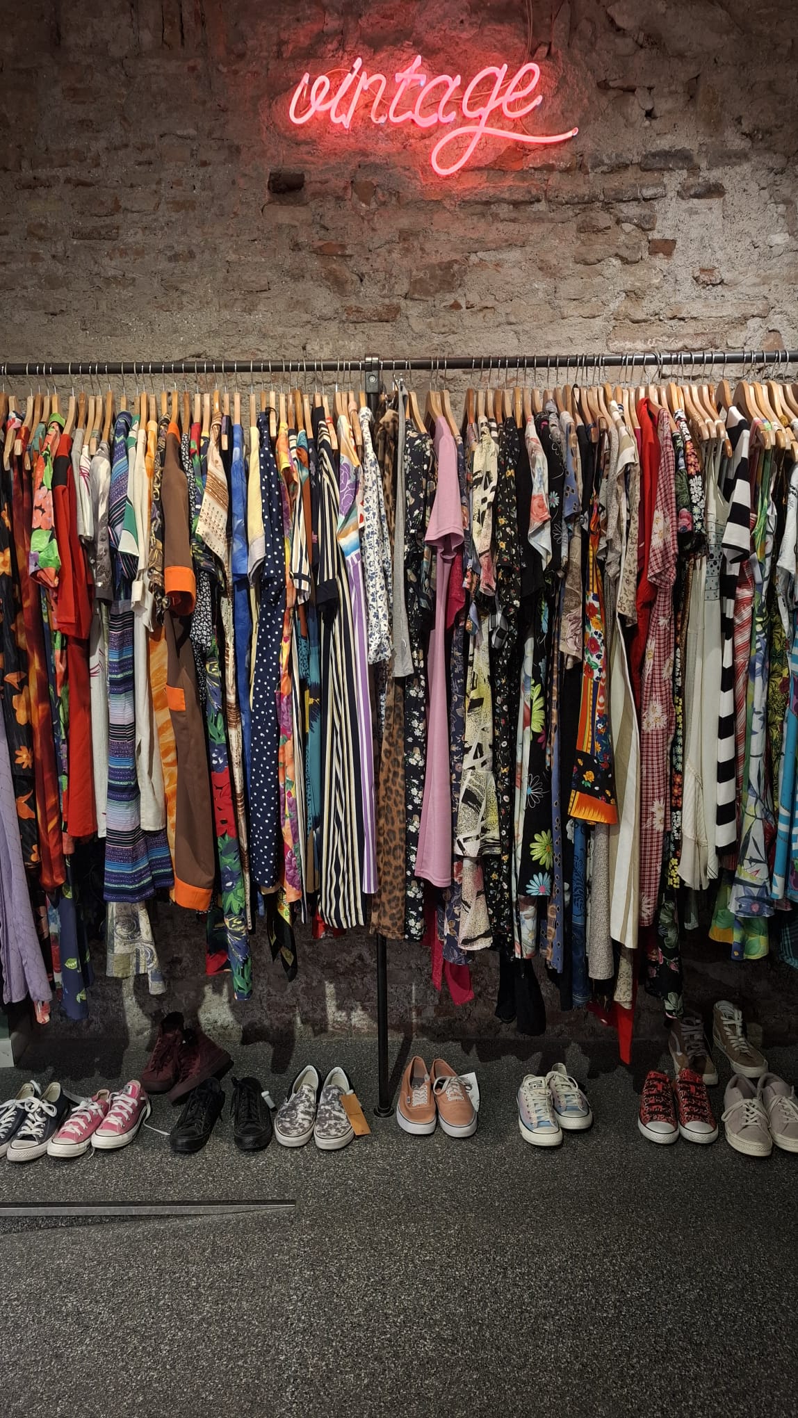 Kleiderstange mit bunter Vintage-Kleidung und Schuhen unter einem Vintage-Neonschild bei Península Vintage in Málaga.